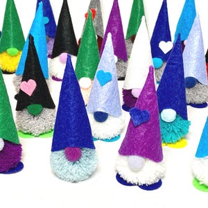 May include: A collection of colorful gnome figurines with cone-shaped hats in various colors, including green, blue, purple, and black. Each gnome has a fluffy beard and a small heart or white heart-shaped accent on the hat. The gnomes are standing on a white surface.