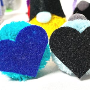 May include: Close-up of handmade gnome ornaments with colorful pom-pom bodies and felt heart accents. The gnomes have various colored hats, including green, black, and yellow. The hearts are blue and black.