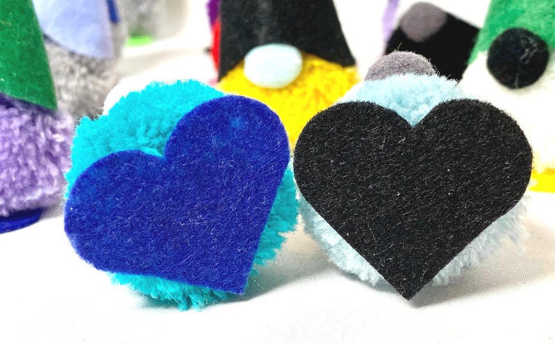 May include: Close-up of handmade gnome ornaments with colorful pom-pom bodies and felt heart accents. The gnomes have various colored hats, including green, black, and yellow. The hearts are blue and black.