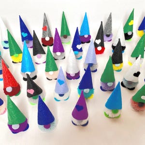 May include: A collection of colorful gnome figurines with cone-shaped hats in various colors, including red, green, blue, and gray. Each gnome has a fluffy white beard and a heart or other shape on the hat. The gnomes are arranged on a white surface.