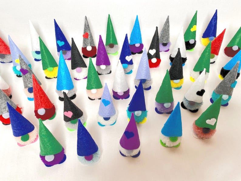 May include: A collection of colorful gnome figurines with cone-shaped hats in various colors, including red, green, blue, and gray. Each gnome has a fluffy white beard and a heart or other shape on the hat. The gnomes are arranged on a white surface.
