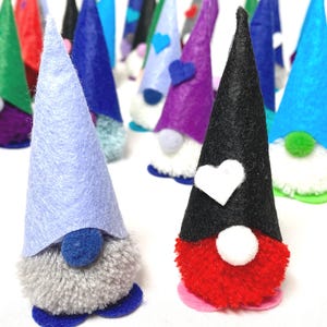 May include: A collection of handmade gnome figurines with felt cone hats in various colors, including blue, green, purple, and black. Each gnome has a fluffy pom-pom beard and a small base. Some hats feature heart-shaped accents.