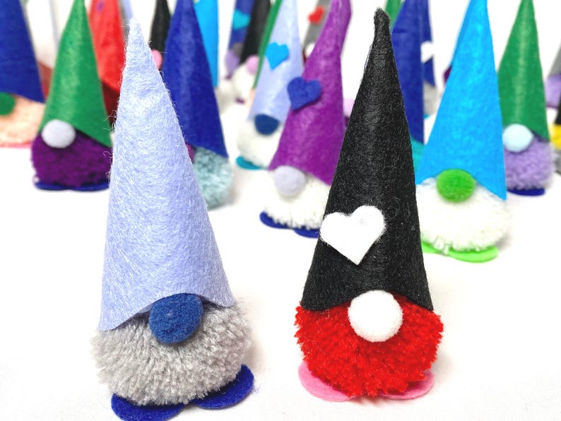 May include: A collection of handmade gnome figurines with felt cone hats in various colors, including blue, green, purple, and black. Each gnome has a fluffy pom-pom beard and a small base. Some hats feature heart-shaped accents.
