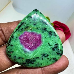 May include: A teardrop-shaped gemstone with a vibrant green base, speckled with black inclusions. The center features a striking, textured magenta-colored core. The stone is held in a hand, showcasing its polished surface and natural beauty.