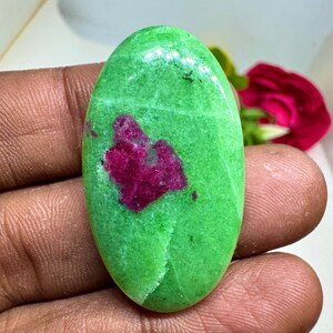 May include: Oval-shaped gemstone featuring a vibrant green base color with a prominent, irregular magenta-colored inclusion. The polished stone is held in a hand, showcasing its smooth surface and natural variations. The gemstone is a unique piece of jewelry.