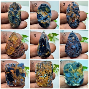May include: A collection of nine polished pietersite gemstones in various shapes, including ovals, teardrops, and rectangles. Each stone displays a unique pattern of deep blue, gold, and brown swirling fibers, numbered one through nine.