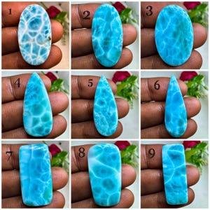 Op de afbeelding: Een collectie van negen gepolijste Larimar edelstenen in diverse vormen, waaronder ovalen, druppels en rechthoeken. Elke steen vertoont een levendige hemelsblauwe kleur met witte, webachtige patronen, genummerd van één tot en met negen.