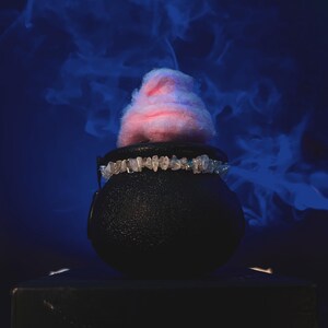 May include: A black cauldron with a textured surface, topped with pink and purple cotton candy. A decorative band with clear stones encircles the cauldron. The background is a dark blue with wisps of blue smoke.