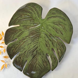 May include: A ceramic leaf-shaped decorative plate with a glossy green finish and brown veining. The plate has a gold-colored trim and is designed to resemble a Monstera leaf. The plate is a decorative item for the home.