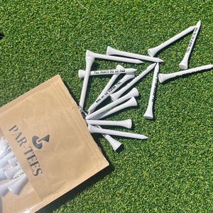 以下が含まれることがあります： 「PAR-TEES」と印刷された茶色の紙袋。袋は部分的に開いており、「TIL DEATH DO US PAR」と印刷された白いゴルフティーがこぼれています。ティーは緑の人工芝の上にあります。