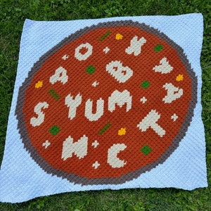 May include: A crocheted blanket with a pizza design. The pizza has the words "S'YUM M'C" in white yarn on a brown background. The pizza is surrounded by a blue border.