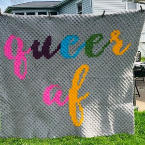 May include: A gray crocheted blanket with the words "queer af" spelled out in colorful yarn.