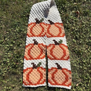 May include: A handmade crochet scarf featuring a repeating pumpkin design. The scarf has a light gray background with orange, tan, and brown pumpkins. The scarf is lying on a bed of green grass.