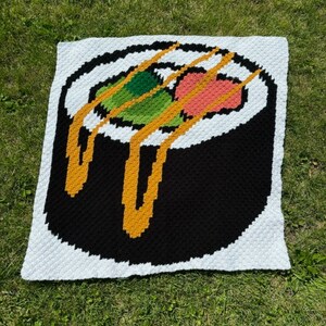 Puede incluir: Una manta de ganchillo con un rollo de sushi colorido con hilo amarillo, verde, rosa y negro. La manta es blanca con un borde azul claro.