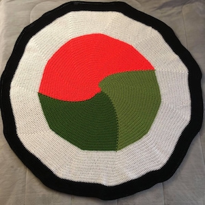 Op de afbeelding: Een rond gebreid vloerkleed met een zwarte rand en een veelkleurig midden. Het midden is verdeeld in drie secties: rood, groen en donkergroen.