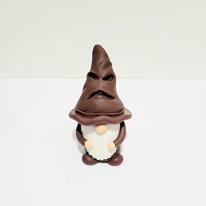 May include: A whimsical brown gnome figurine with a pointed hat and a long white beard. The hat has a face-like design. The gnome has a round body and small arms. The figurine is on a white surface.
