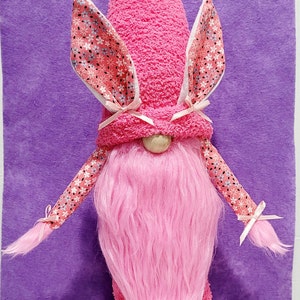 May include: A pink gnome with a pink fuzzy beard and a pink hat with bunny ears. The hat has a floral pattern and the gnome is wearing pink slippers.