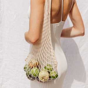 May include: A person wearing a white dress carries a white mesh bag filled with green and yellow fruit.