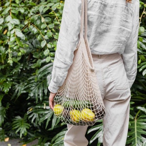 以下が含まれることがあります： 白いメッシュバッグに緑のアボカドと黄色のレモンが詰まっている人を写した写真。バッグは肩にかけられており、その人は白いシャツと白いズボンを着用している。