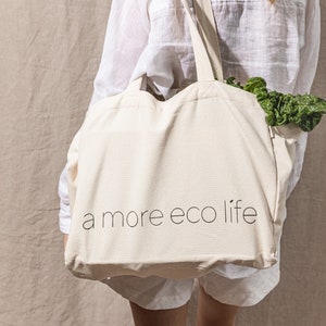 May include: A person carrying a white canvas tote bag with the text "a more eco life" printed on it. The bag is filled with leafy green vegetables.