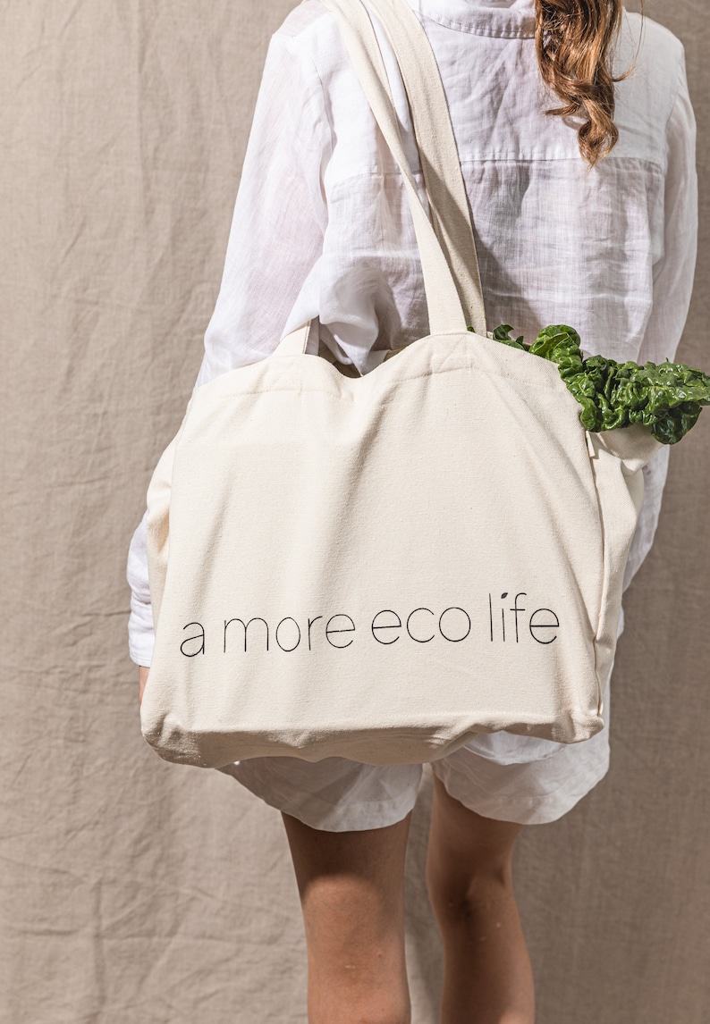 May include: A person carrying a white canvas tote bag with the text "a more eco life" printed on it. The bag is filled with leafy green vegetables.