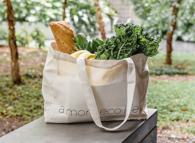 May include: A white canvas tote bag with the text "amor ecolife" printed on the front. The bag is filled with groceries, including a loaf of bread, bananas, and leafy green vegetables.