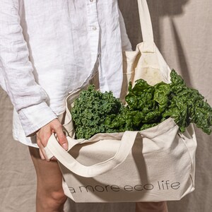May include: A person holding a white tote bag with the text "a more eco life" printed on it. The bag is filled with green leafy vegetables, such as kale and spinach.