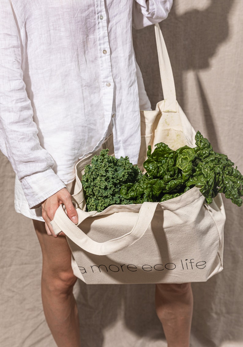 May include: A person holding a white tote bag with the text "a more eco life" printed on it. The bag is filled with green leafy vegetables, such as kale and spinach.