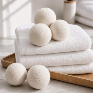 May include: A collection of white wool dryer balls arranged on a stack of folded white towels. The balls are spherical and have a textured surface. A wooden tray and a white soap dispenser are also visible in the background. The scene is brightly lit.