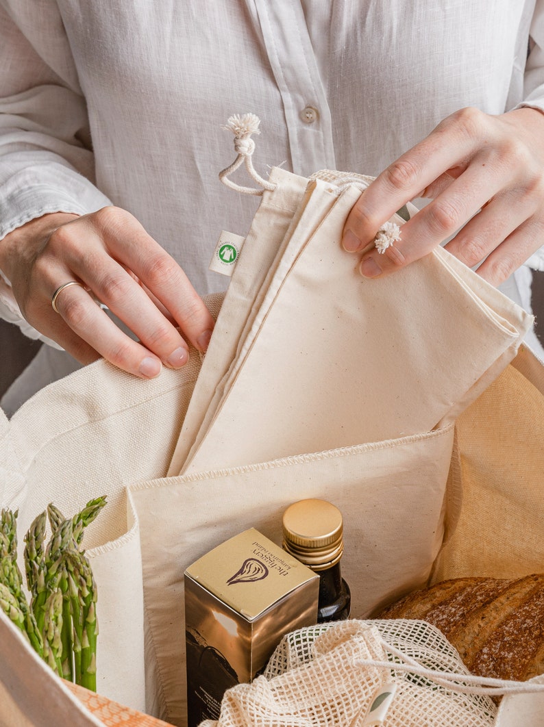 May include: A white canvas tote bag with multiple pockets filled with groceries. The bag contains a bottle of olive oil, a box of The Hersey Extra Virgin Olive Oil, a bunch of asparagus, and a loaf of bread in a mesh bag.