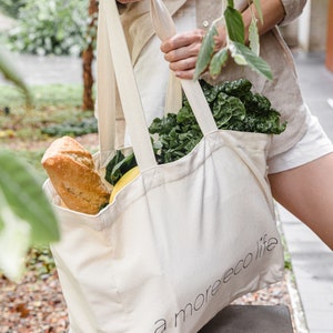May include: A white canvas tote bag with the text "a more eco life" printed on the side. The bag is filled with groceries, including a loaf of bread and leafy green vegetables.