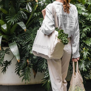 May include: A person carrying a beige tote bag with the text "a more eco life" printed on it. The bag is filled with leafy green vegetables. The person is also carrying a white mesh bag with more vegetables.
