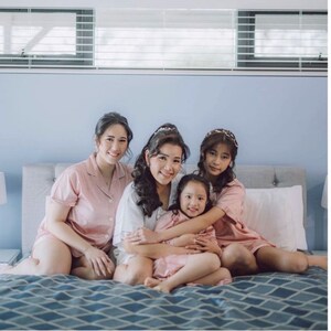 Puede incluir: Cuatro mujeres en pijama de sat&eacute;n rosa est&aacute;n sentadas en una cama. La mujer del medio lleva una camisa blanca y tiene los brazos alrededor de una ni&ntilde;a peque&ntilde;a. La ni&ntilde;a tambi&eacute;n lleva una parte superior de pijama de sat&eacute;n rosa. Las mujeres sonr&iacute;en y miran a la c&aacute;mara.