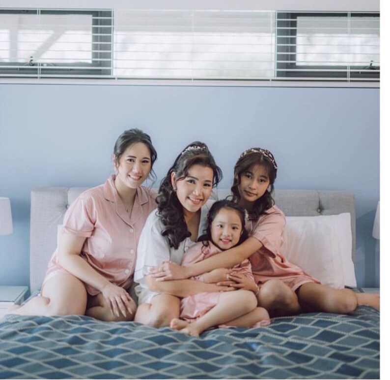 Puede incluir: Cuatro mujeres en pijama de sat&eacute;n rosa est&aacute;n sentadas en una cama. La mujer del medio lleva una camisa blanca y tiene los brazos alrededor de una ni&ntilde;a peque&ntilde;a. La ni&ntilde;a tambi&eacute;n lleva una parte superior de pijama de sat&eacute;n rosa. Las mujeres sonr&iacute;en y miran a la c&aacute;mara.