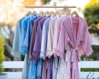 May include: A row of eight robes hanging on a wooden rack. The robes are in various shades of blue, purple, and pink. Each robe has a lace trim around the edges.