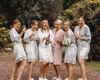 May include: Six women in matching light blue satin robes with lace trim, smiling and holding champagne flutes. They are standing in a garden setting with green foliage and purple flowers.