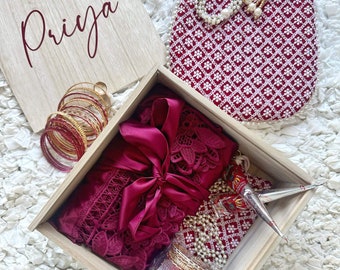 Puede incluir: Una caja de madera con la palabra "Priya" en la tapa. La caja está abierta y contiene una bata de satén burdeos con ribete de encaje, una pequeña bolsa roja, una cadena de perlas blancas y varios brazaletes dorados.