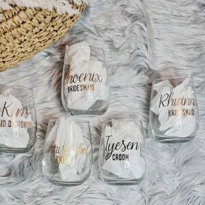 Op de afbeelding: Vijf heldere glazen wijnglazen met gouden letters. De glazen zijn gepersonaliseerd met de namen "Kim Maid of Honor", "Christabel Bride", "Tyesen Groom", "Phoenix Bridesmaid" en "Rhianna Bridesmaid".