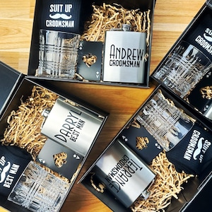 May include: Four gift boxes with black lids and brown shredded paper inside. Each box contains a black and white patterned sock, a clear glass, a silver flask, and a black card with a playing card design. The flasks are engraved with the names "Andrew", "Darryl", "Terrance", and "Suit Up Groomsmans".