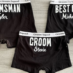 May include: Three pairs of black boxer shorts with white elastic waistbands. The waistbands have the text "Groom Squad" printed on them. The boxer shorts have white text printed on the back. The text on the boxer shorts reads "Groom", "Best Man", and "Groomsman". The names "Stevie", "Michael", and "Tyler" are printed below the titles.