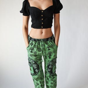 May include: A black crop top with gold buttons and short sleeves is paired with green patterned pants. The pants feature a black and green Celtic knot design. The outfit is completed with white sneakers.