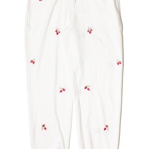 May include: White sweatpants with an elastic waistband and drawstring. The pants are adorned with embroidered red strawberries and green leaves scattered across the fabric. The casual pants have two pockets and a relaxed fit.
