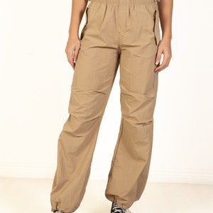 May include: Tan cargo trousers with an elasticated waistband and drawstring details. The trousers have pockets and are gathered at the ankles. The model is wearing black and white trainers.