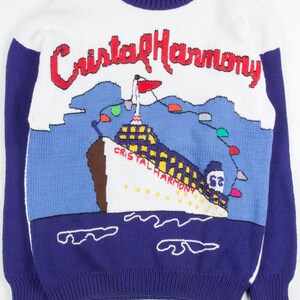 Könnte beinhalten: Ein weißer, blauer und lila gestrickter Pullover mit einem Cartoon-Bild eines Kreuzfahrtschiffs mit dem Text "Cristal Harmony" darauf. Der Pullover hat einen blau-weiß gestreiften Besatz an Hals und Ärmeln.