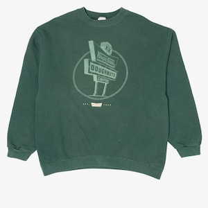 May include: Dark green sweatshirt featuring a circular graphic with the text "Krispy Kreme Doughnuts Coffee". The design includes a stylized sign with the brand's logo. The sweatshirt has long sleeves and a crew neck.
