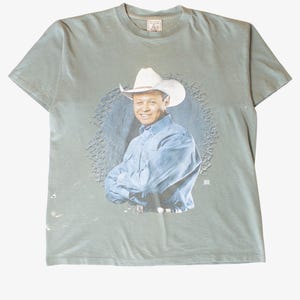 May include: Sage green t-shirt featuring a portrait of a man wearing a cowboy hat and a blue button-down shirt. The portrait is set against a blue circular background with a decorative border. The t-shirt has a crew neck and short sleeves.
