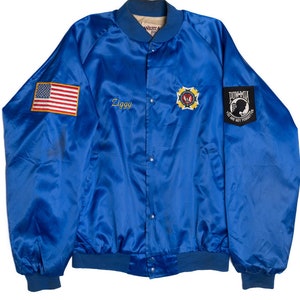 Może przedstawiać: Niebieska satynowa kurtka bomber zapinana na zamek błyskawiczny i zatrzaski. Kurtka ma naszywkę z flagą amerykańską na lewym rękawie, złotą haftowaną naszywkę z gwiazdą i orłem po prawej stronie oraz czarną naszywkę z napisem "POW-MIA You Are Not Forgotten" na prawym rękawie. Kurtka ma również wyhaftowane imię "Ziggy" po lewej stronie klatki piersiowej.