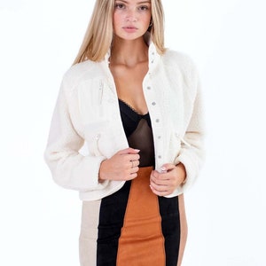 Op de afbeelding: Een wit nep bontjasje met een rits en knoopsluiting. Het jasje is kort en heeft een ontspannen pasvorm. Het model draagt een zwart kanten topje en een bruin en zwart mini-rokje.