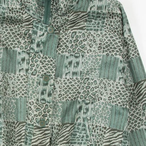 Könnte beinhalten: Eine grün-grau gemusterte Jacke mit hohem Kragen und Reißverschluss. Die Jacke hat eine Mischung aus Tiermustern, darunter Leopardenflecken und Zebrastreifen, und ist vorne mit Knöpfen versehen.