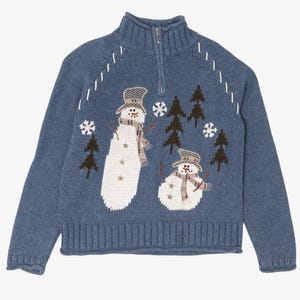 May include: A blue knit sweater featuring a quarter-zip collar and a winter scene. The design includes two white snowmen wearing plaid hats and scarves, surrounded by dark brown pine trees and white embroidered snowflakes on the chest.